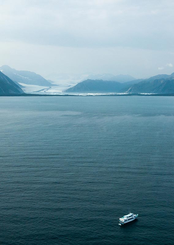 A boat cruises on the sea between forested mountains and a glacier in the distance.