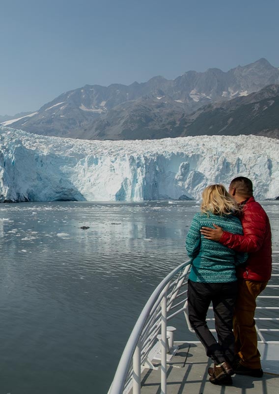 Two people stand at a boat railing, looking towards a tidewater glacier.