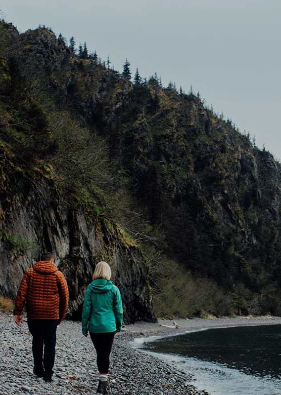 Couple walks along pebbled beach of tranquil Fox Island as mountains jut up around them