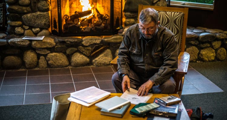 A man sits at a table of historical documents in front of a fireplace.