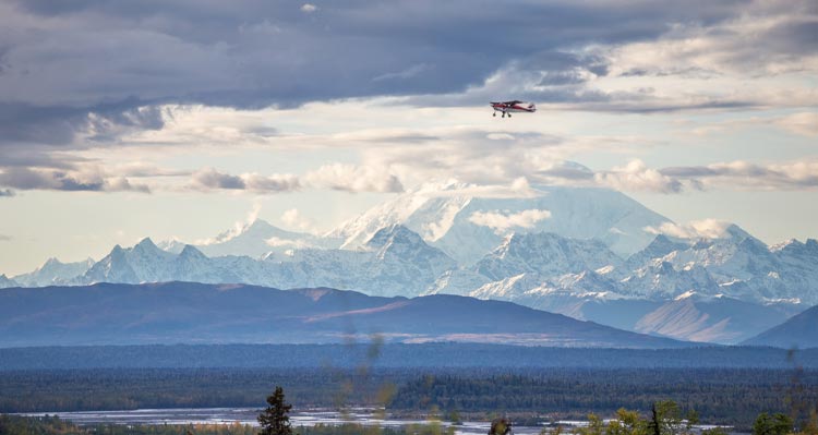 A small red plane flies above forests with mountains in the background.