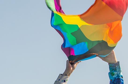 Rainbow flag held in two hands before blue sky