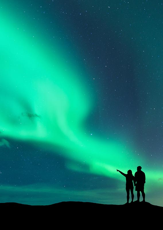 Two people in silhouette look out at a green aurora in the night sky.
