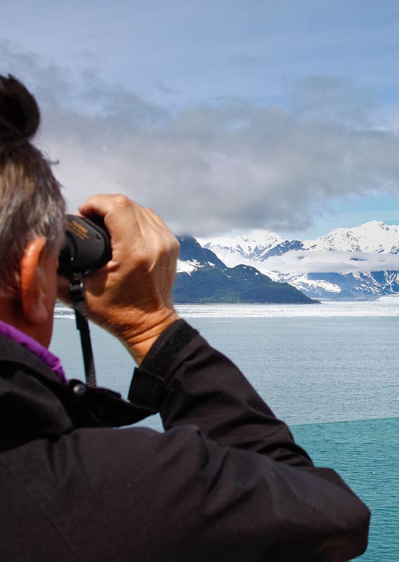 A man from behind looks through binoculars to an ice sheet from the outside of a tour boat.