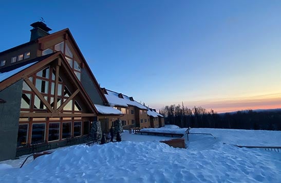 A large wooden lodge covered in snow at sunrise.