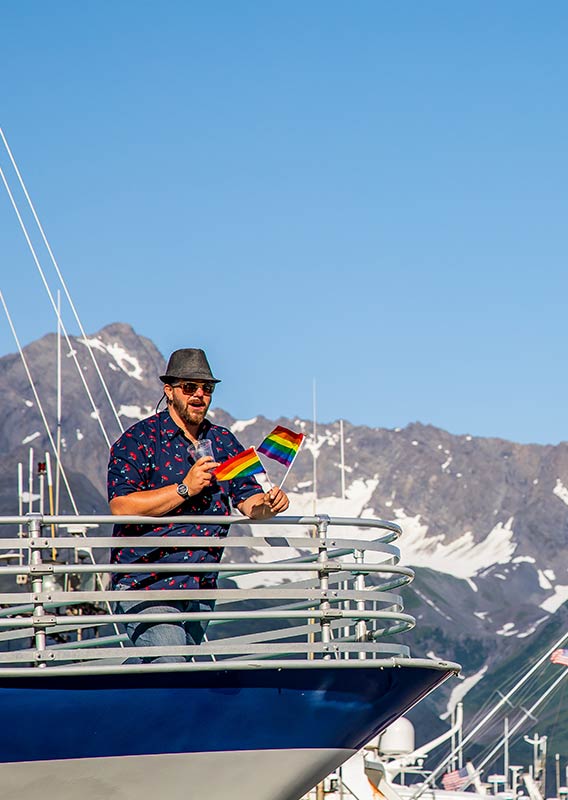 A man stands on the edge of a boat holding small pride flags.