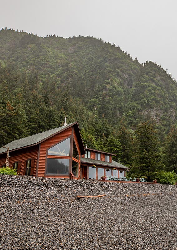 A wooden lodge at the top of a rocky beach below a mist forested, mountainside.